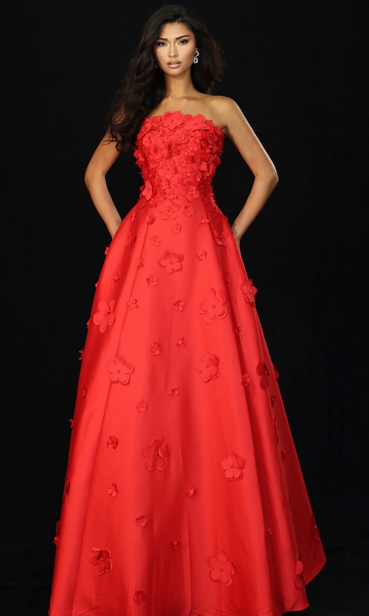 A woman poses against a black background in the Jovani 48444 long prom dress, featuring a strapless red A-line silhouette adorned with three-dimensional floral appliqués.